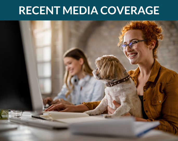 A smiling woman sits at an office computer while holding a dog in her lap.